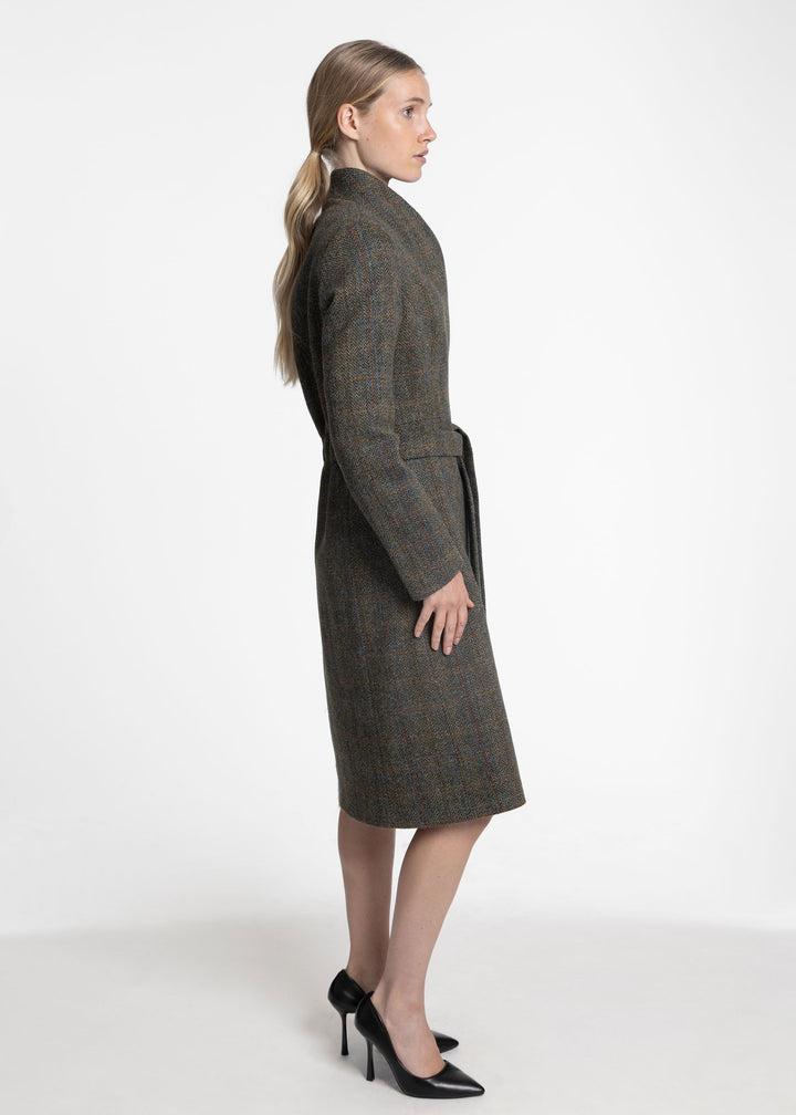 A woman in profile wearing a knee-length green plaid coat with a matching waist tie, paired with black high heels, against a plain white studio background.