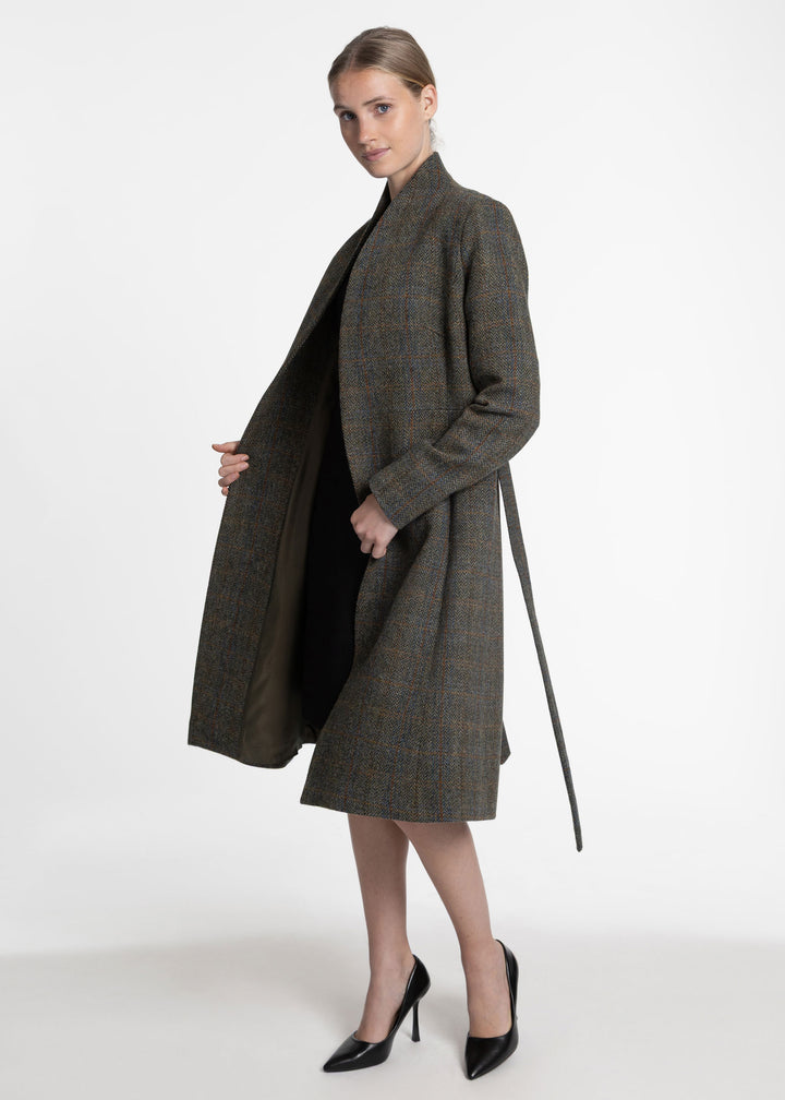 A woman models a mid-length, open-front green coat with a self-tie belt in a dark red and blue plaid pattern, styled over a black dress and black pointed-toe heels against a plain white studio background.