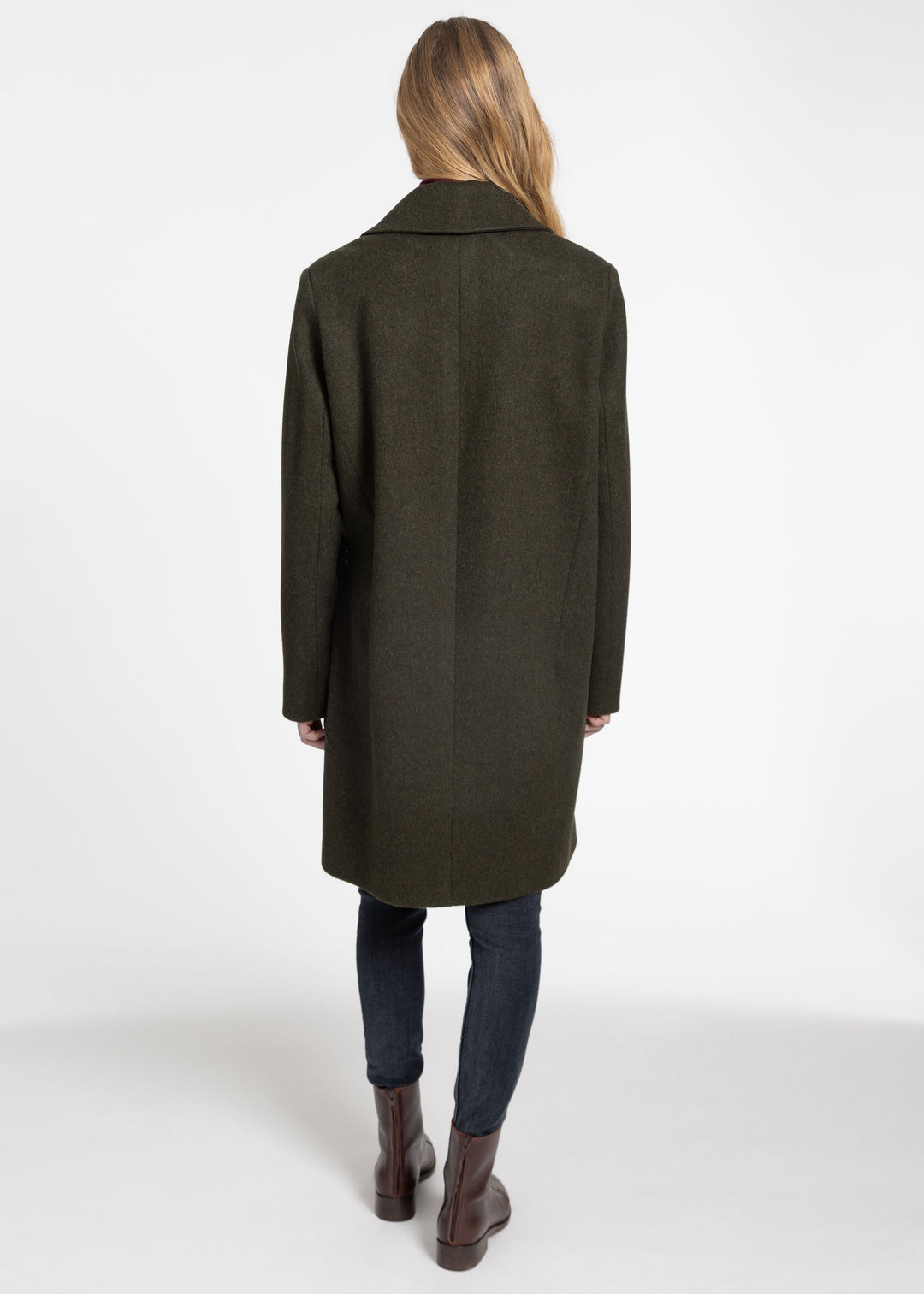 A woman viewed from behind wearing a dark olive green wool coat, dark blue jeans, and brown leather ankle boots against a plain white studio background.