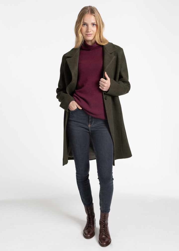 A woman modeling a dark olive green mid-length coat over a burgundy turtleneck sweater, dark blue skinny jeans, and brown leather ankle boots, photographed against a seamless white studio background.
