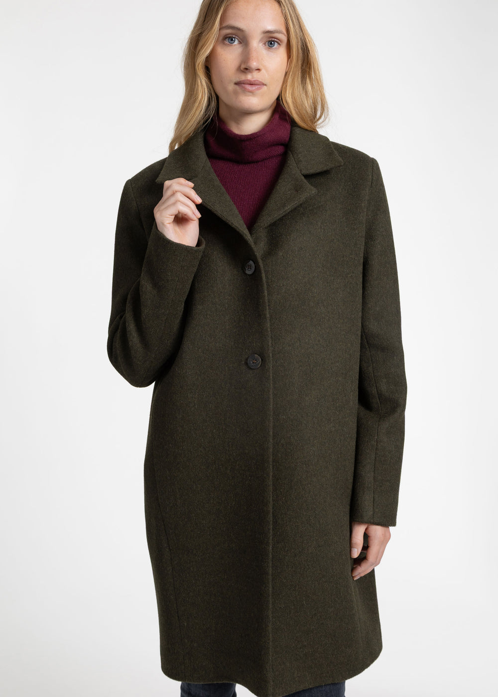 A woman wearing a mid-length olive green wool coat over a burgundy turtleneck sweater, photographed against a plain white studio background.