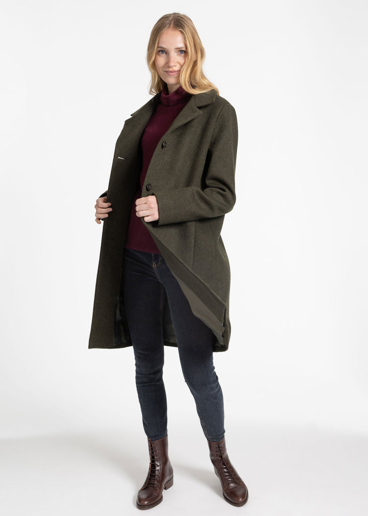 A woman modeling a long olive green coat over a burgundy turtleneck, dark blue jeans, and brown leather ankle boots, photographed against a seamless white studio background.
