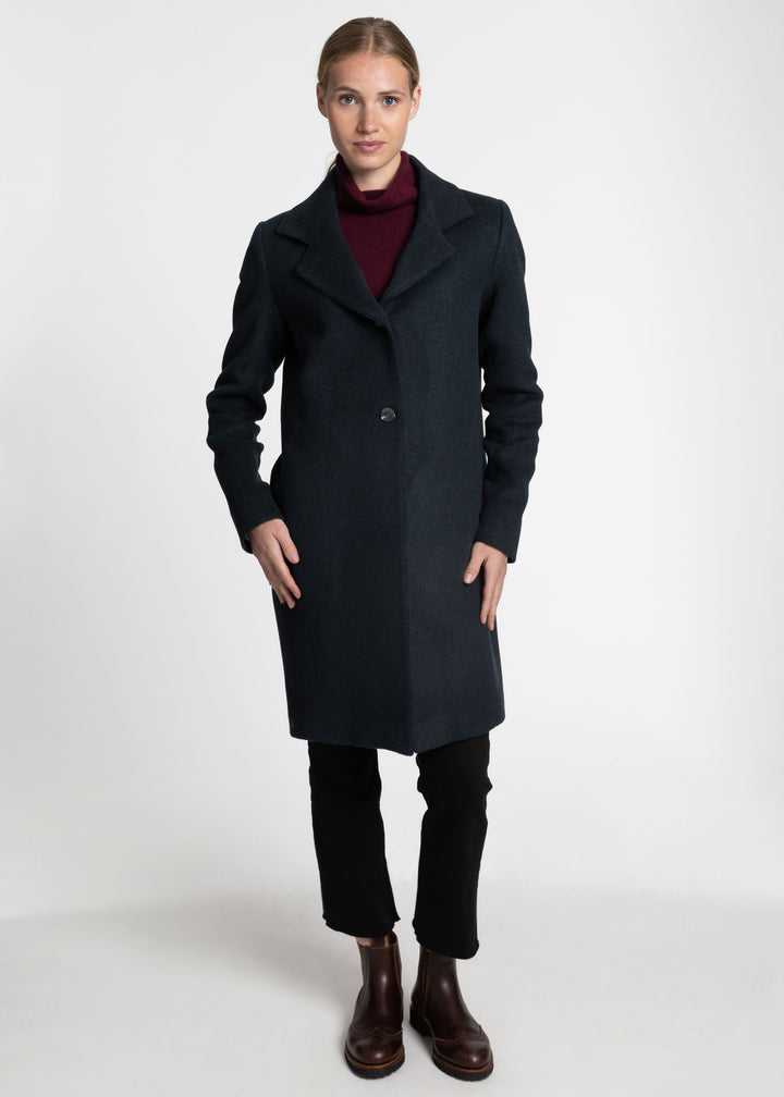 A woman modeling a long dark green wool-blend coat over a burgundy turtleneck, black trousers, and brown leather ankle boots, photographed against a seamless light gray studio background.