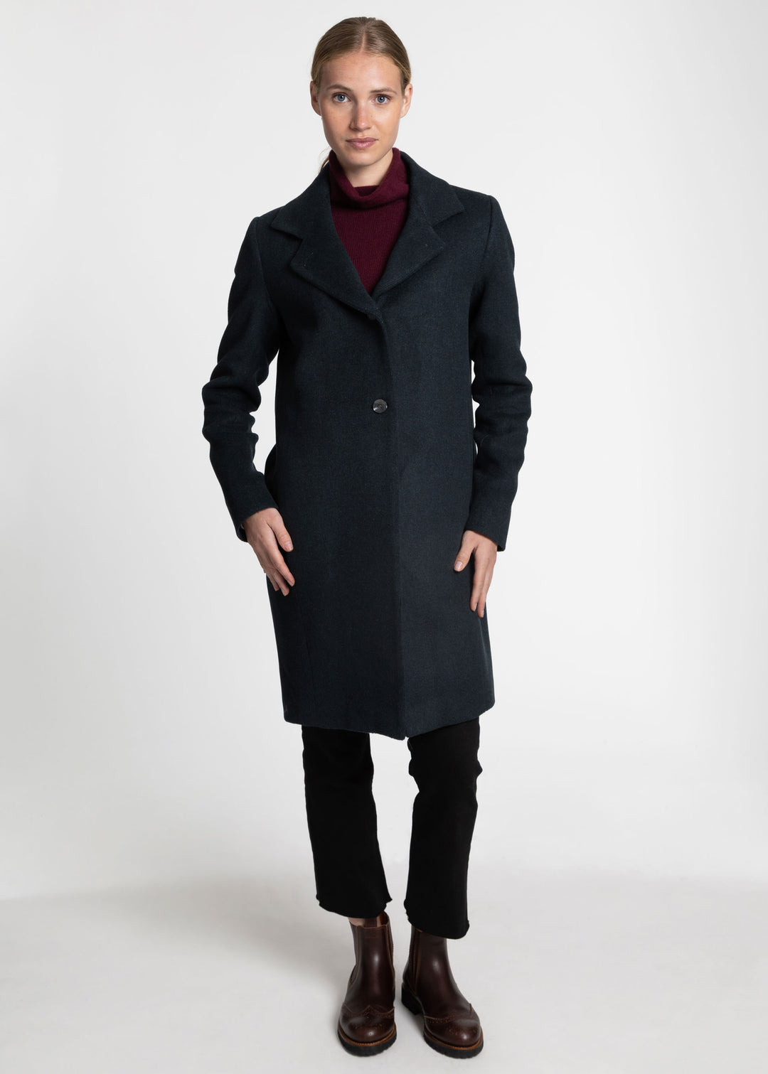 A woman modeling a long dark green wool-blend coat over a burgundy turtleneck, black trousers, and brown leather ankle boots, photographed against a seamless light gray studio background.