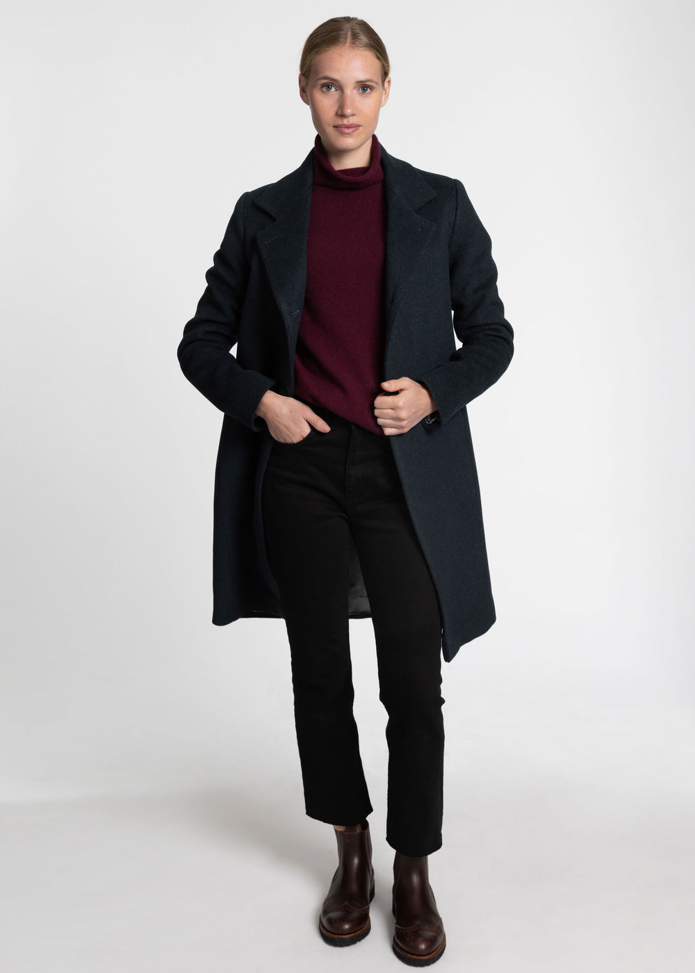 A woman modeling a long dark green coat over a burgundy turtleneck sweater, black trousers, and dark brown ankle boots, photographed against a plain light gray studio background.