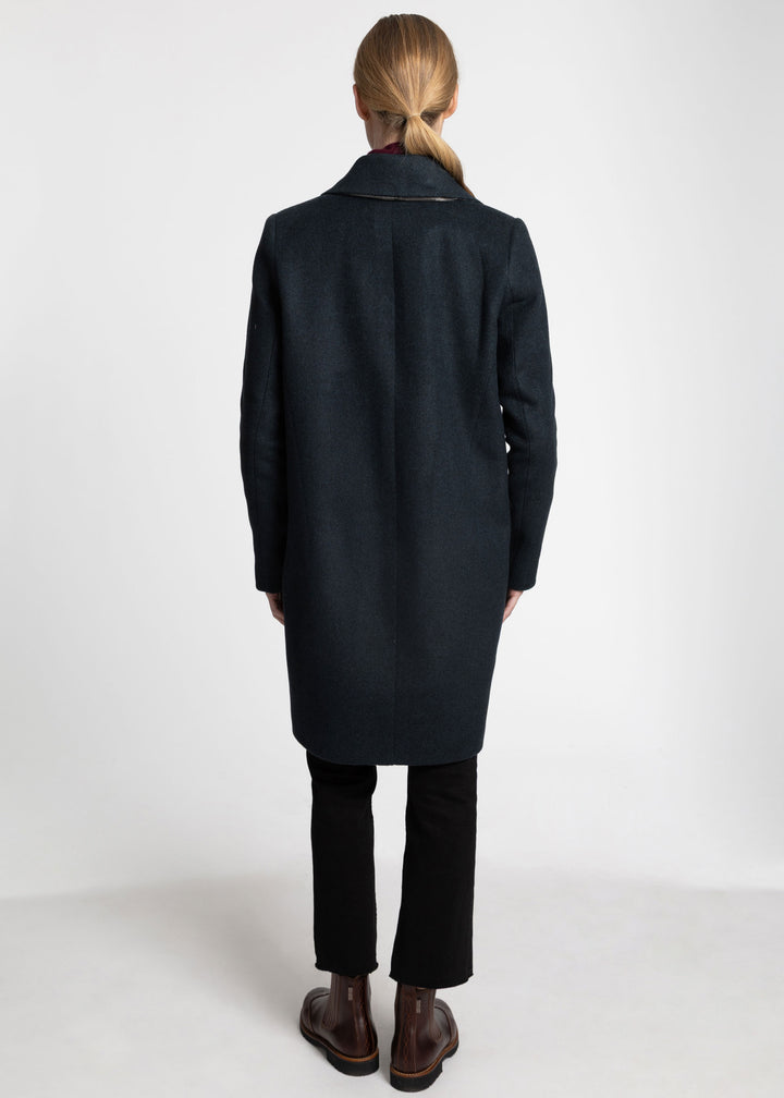 A woman viewed from behind wearing a dark green mid-length coat, black trousers, and brown leather boots against a plain white studio background.