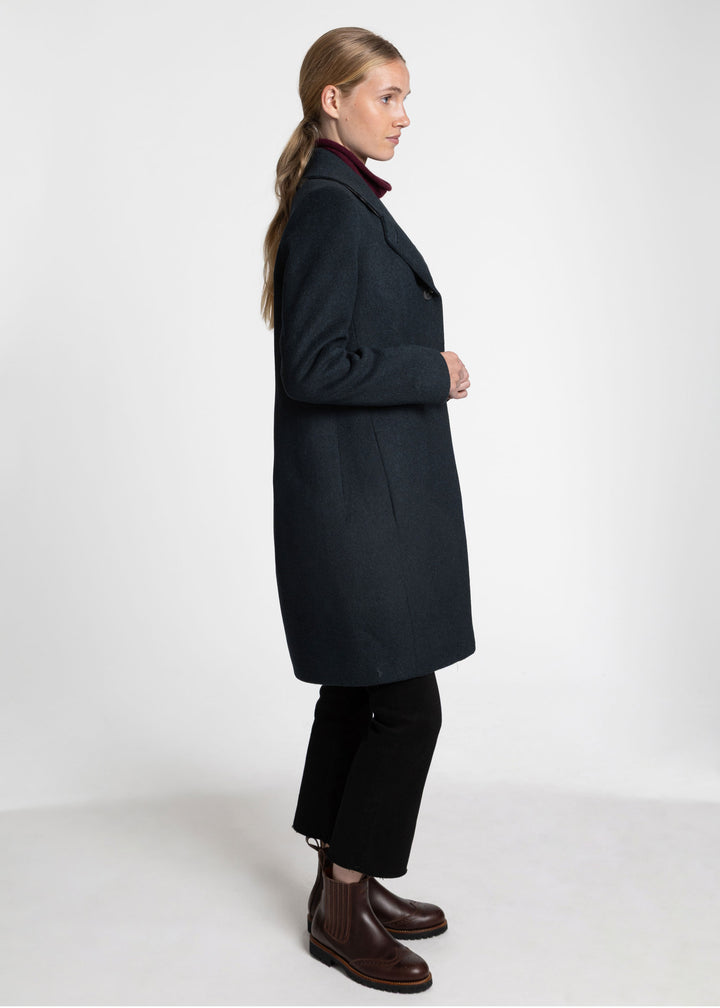 A woman modeling a dark green wool-blend coat, styled with black trousers and brown Chelsea boots, shown in a side profile against a plain white studio background.