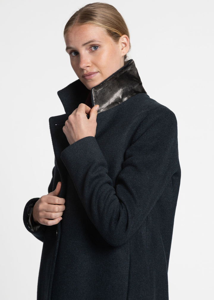 A woman wearing a dark green wool coat with a contrasting silver silk velvet under collar, posing against a light gray studio background.