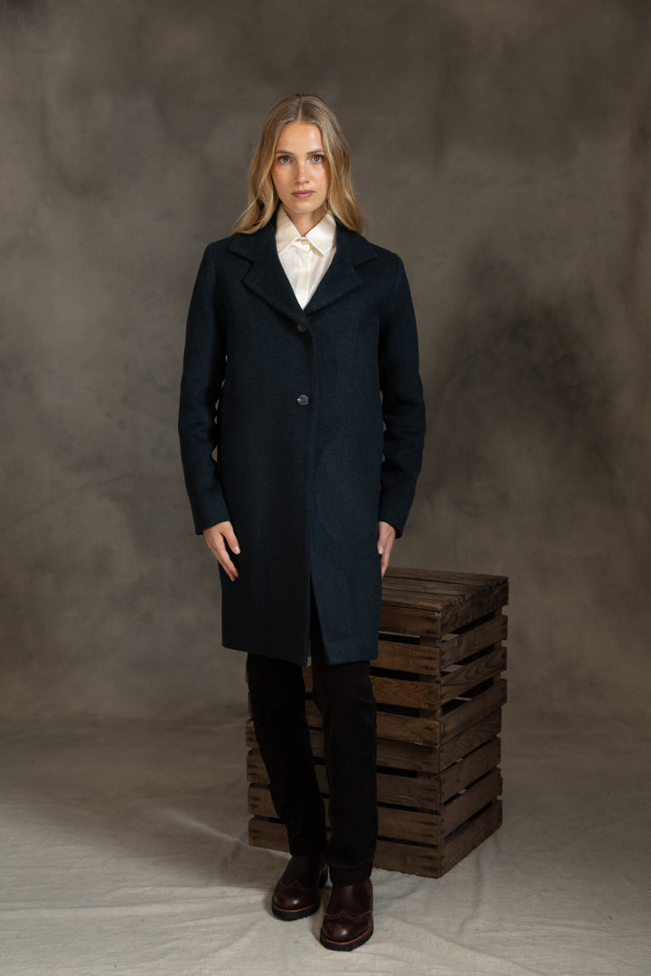 A woman stands in a studio wearing a long dark green coat over a white collared shirt, black pants, and brown shoes, posed beside a stack of wooden crates against a mottled gray backdrop.
