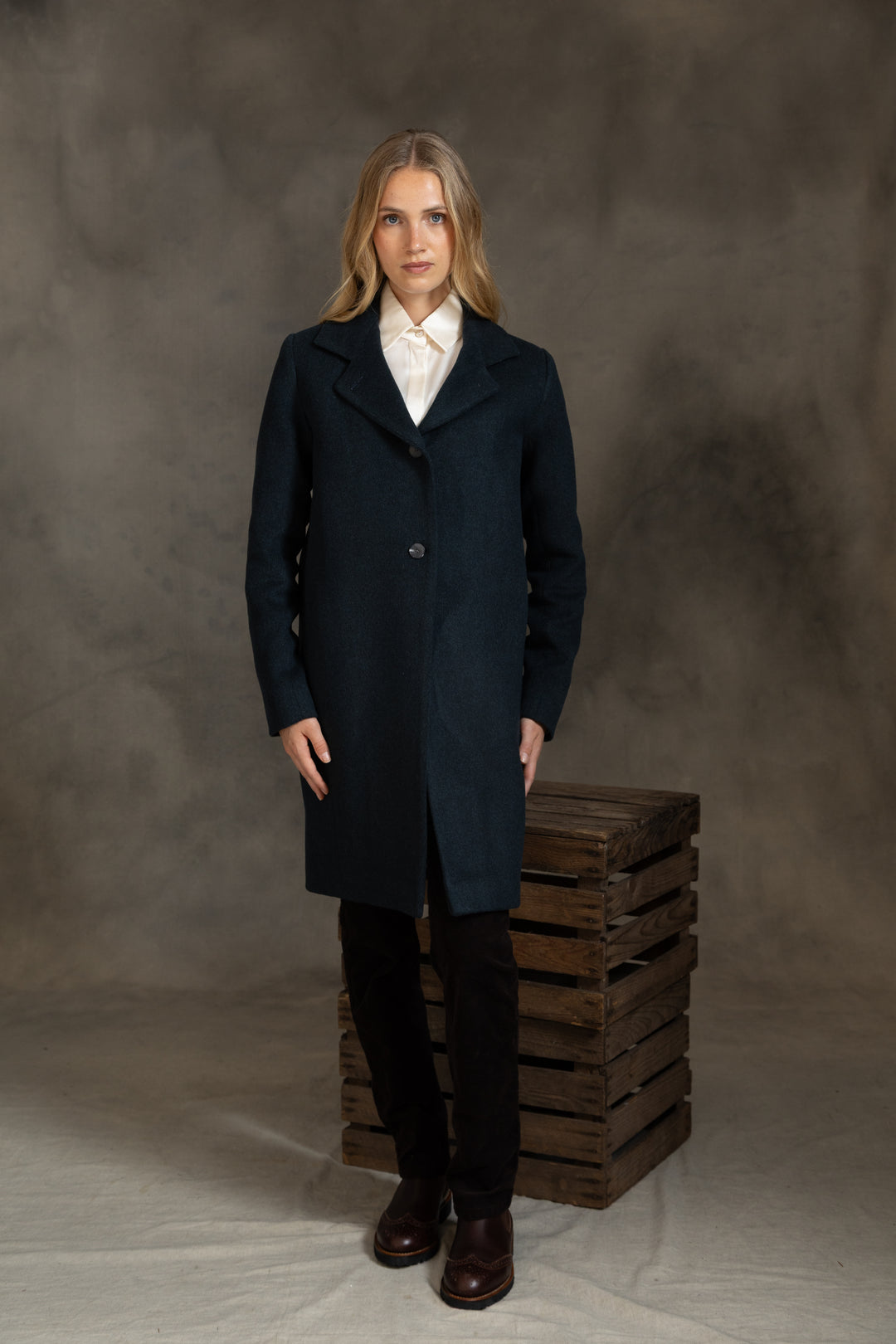 A woman stands in a studio wearing a long dark green coat over a white collared shirt, black pants, and brown shoes, posed beside a stack of wooden crates against a mottled gray backdrop.