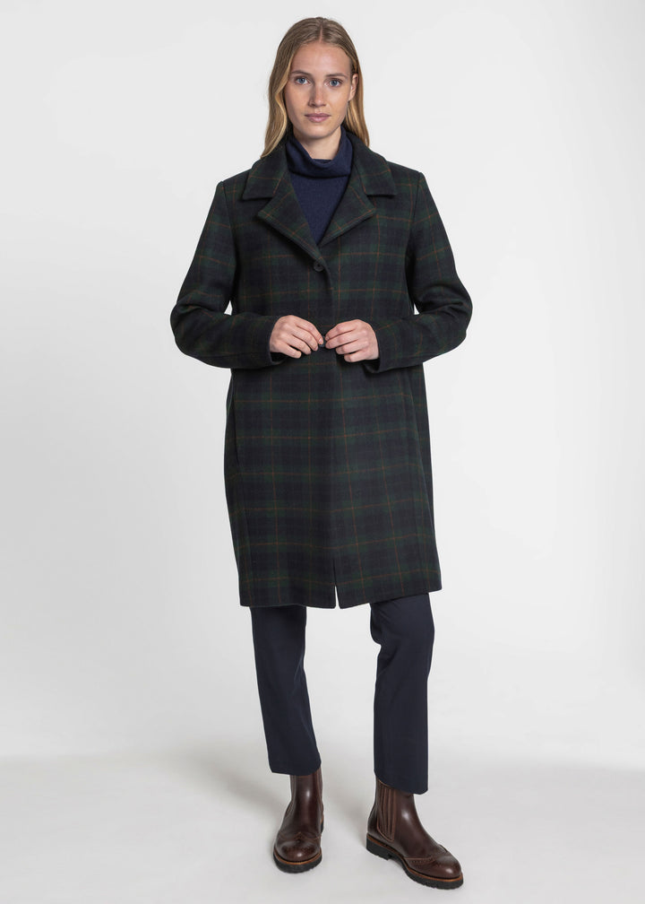 A woman modeling a dark green and black plaid knee-length coat over a navy turtleneck, navy trousers, and brown leather boots, photographed against a plain light gray studio background.