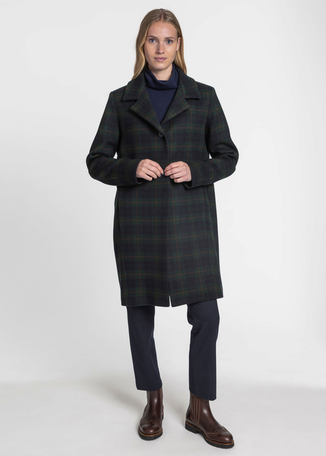 A woman modeling a dark green and black plaid knee-length coat over a navy turtleneck, navy trousers, and brown leather boots, photographed against a plain light gray studio background.
