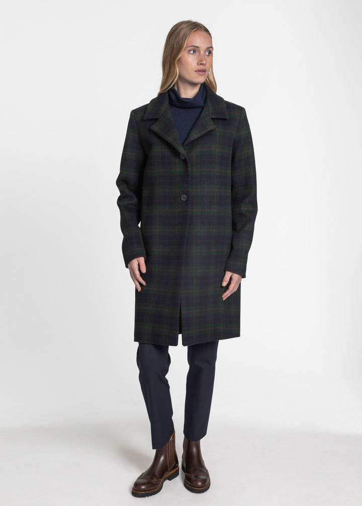 A woman modeling a dark green and black plaid wool coat over a navy turtleneck, navy trousers, and brown leather boots, photographed against a seamless white studio background.