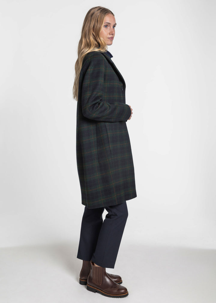 A woman modeling a long dark green and black plaid coat, paired with dark trousers and brown leather boots, photographed against a plain white studio background.