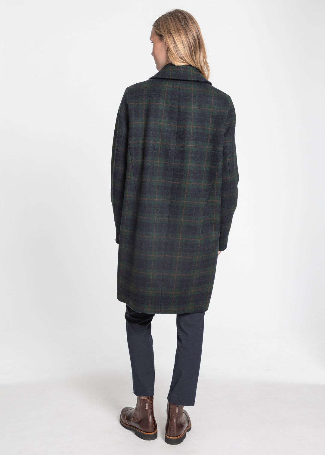 A woman viewed from behind wearing a dark green and black plaid coat, navy trousers, and brown leather boots, standing against a plain white studio background.