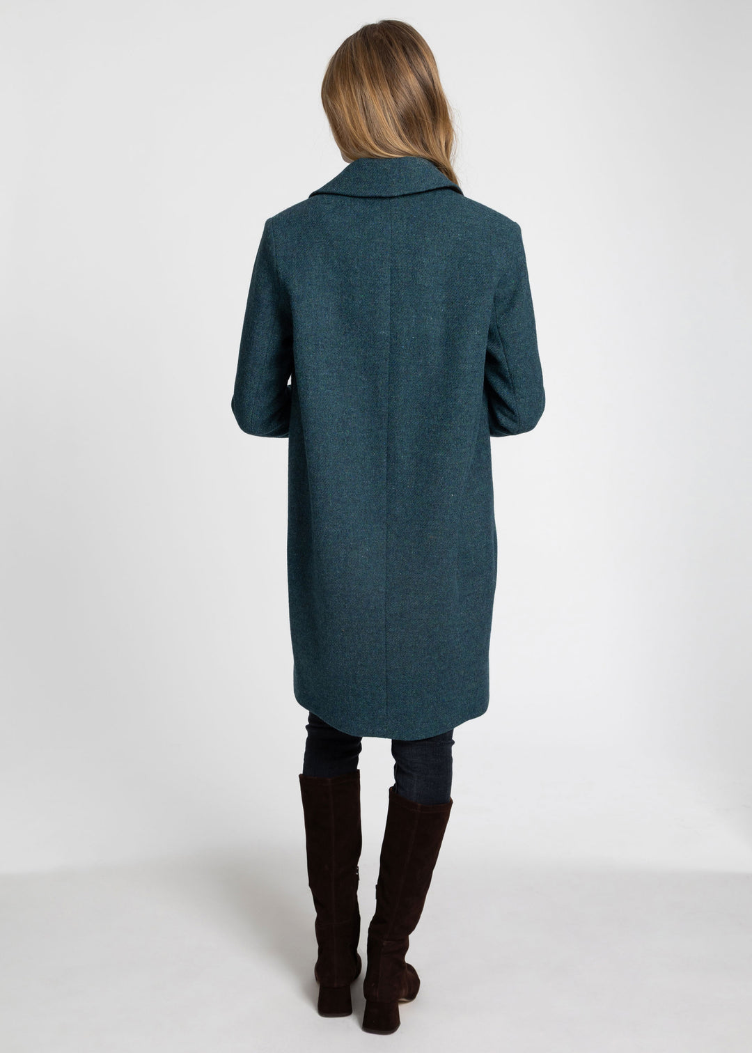 A woman viewed from behind wearing a long teal-blue coat, dark pants, and dark brown knee-high boots against a plain light gray studio background.