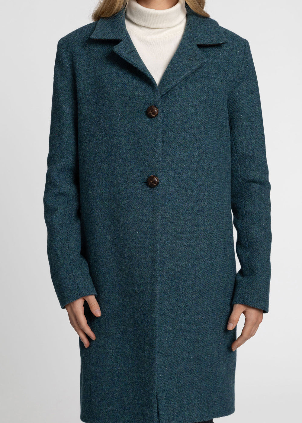 A woman wearing a dark teal blue wool-blend coat over a white turtleneck sweater, shown from the neck down against a light gray studio background.