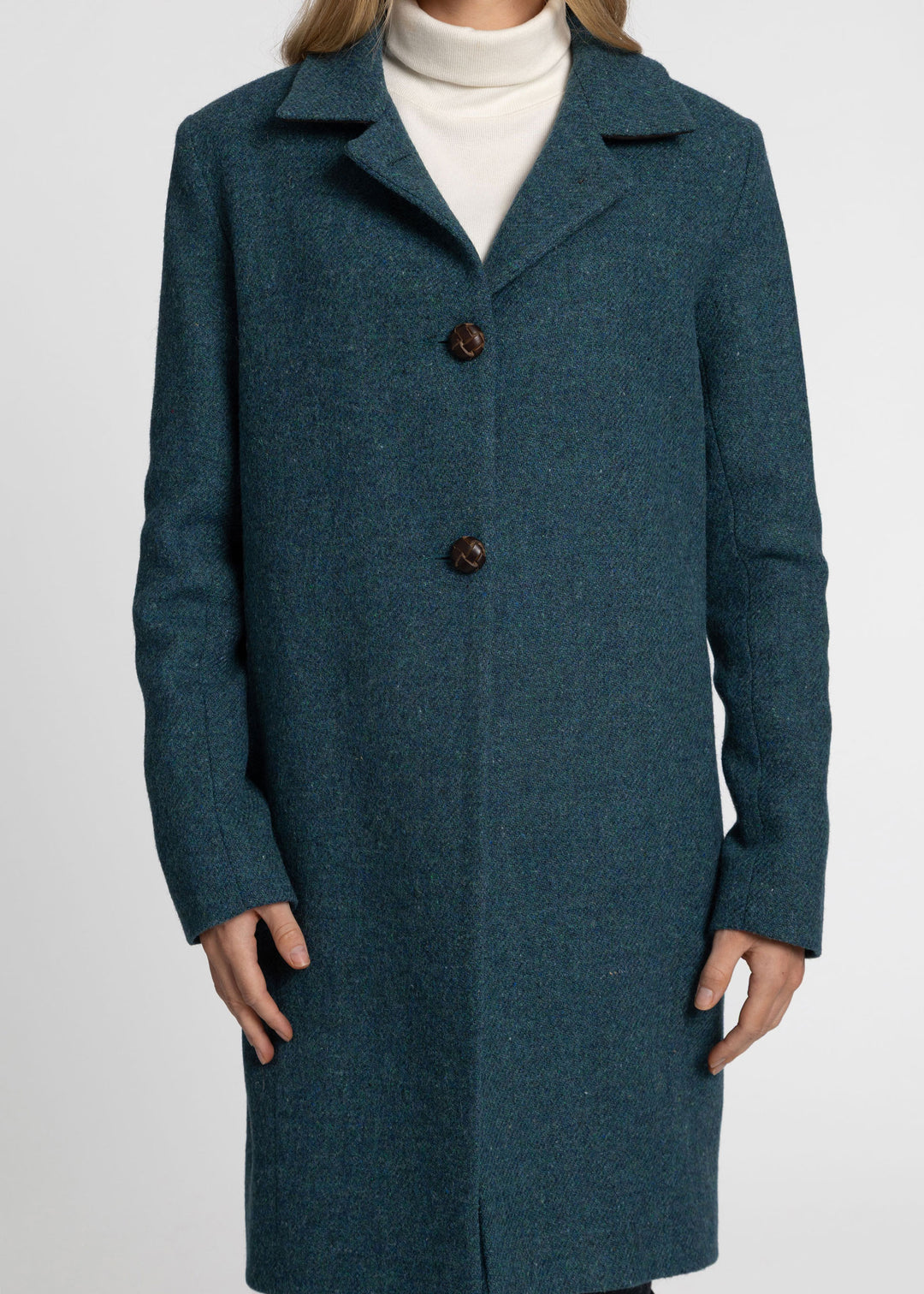 A woman wearing a dark teal blue wool-blend coat over a white turtleneck sweater, shown from the neck down against a light gray studio background.