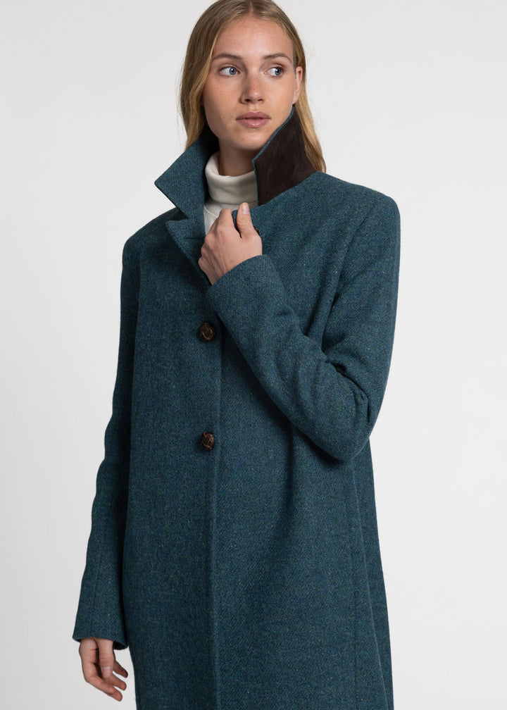 A woman wearing a long teal-blue wool coat with brown suede under the collar, worn over a cream turtleneck sweater photographed against a plain light gray studio background.