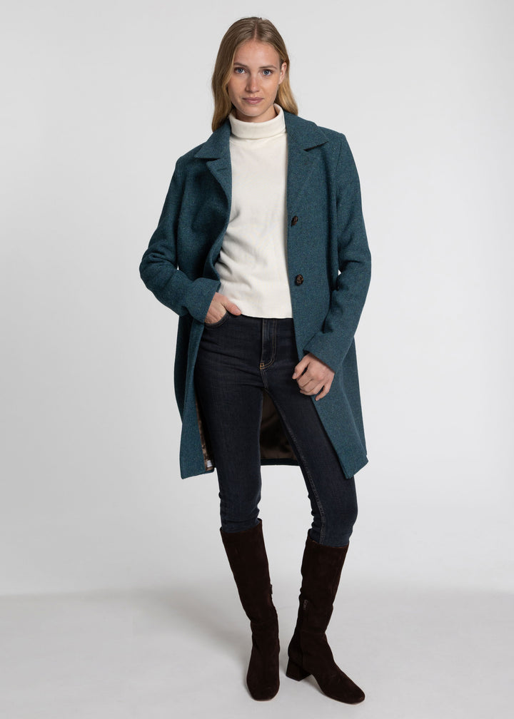 A woman modeling a teal blue-green wool-blend coat over a cream turtleneck, dark skinny jeans, and dark brown knee-high boots, photographed against a light gray studio background.