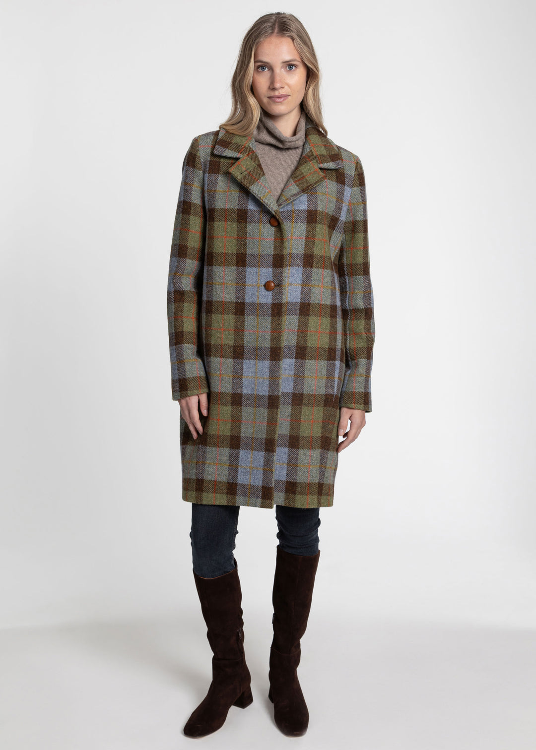 A woman modeling a knee-length plaid coat in muted green, brown, beige, and blue tones, worn over a taupe turtleneck, dark jeans, and dark brown knee-high boots against a plain light gray studio background.