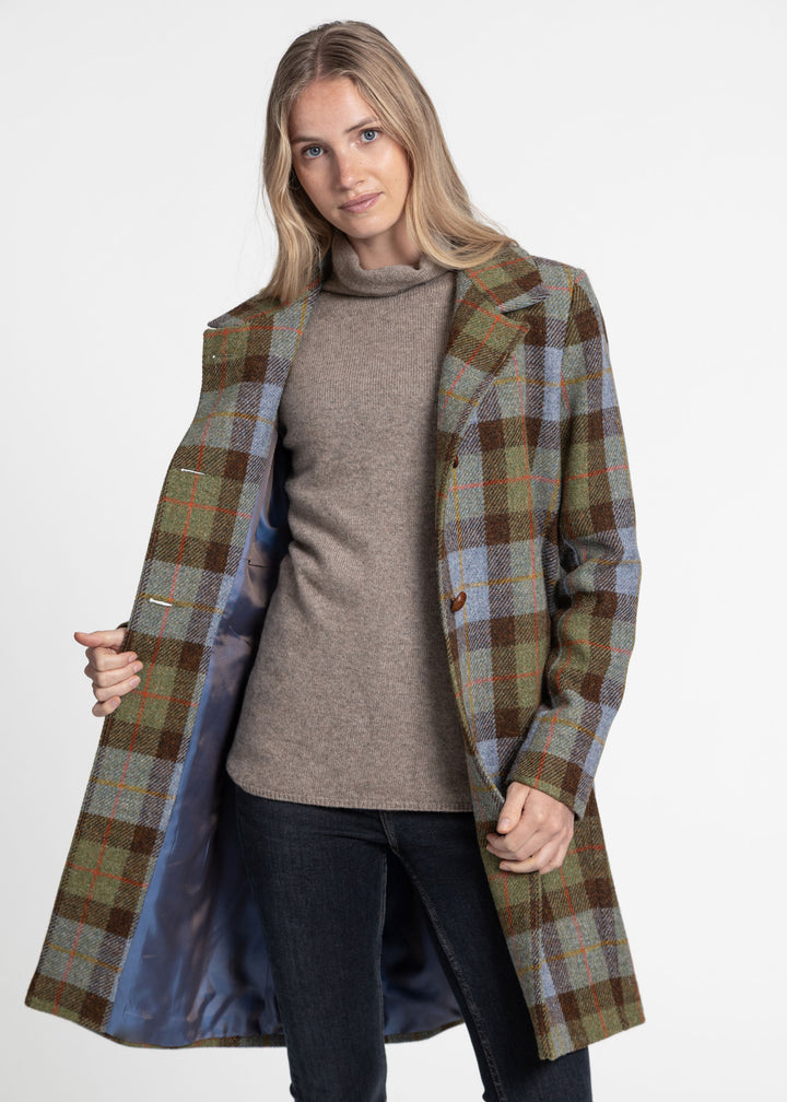 A woman modeling a mid-length plaid coat with a blue lined interior, worn open over a taupe turtleneck sweater and dark jeans, against a plain light gray studio background.
