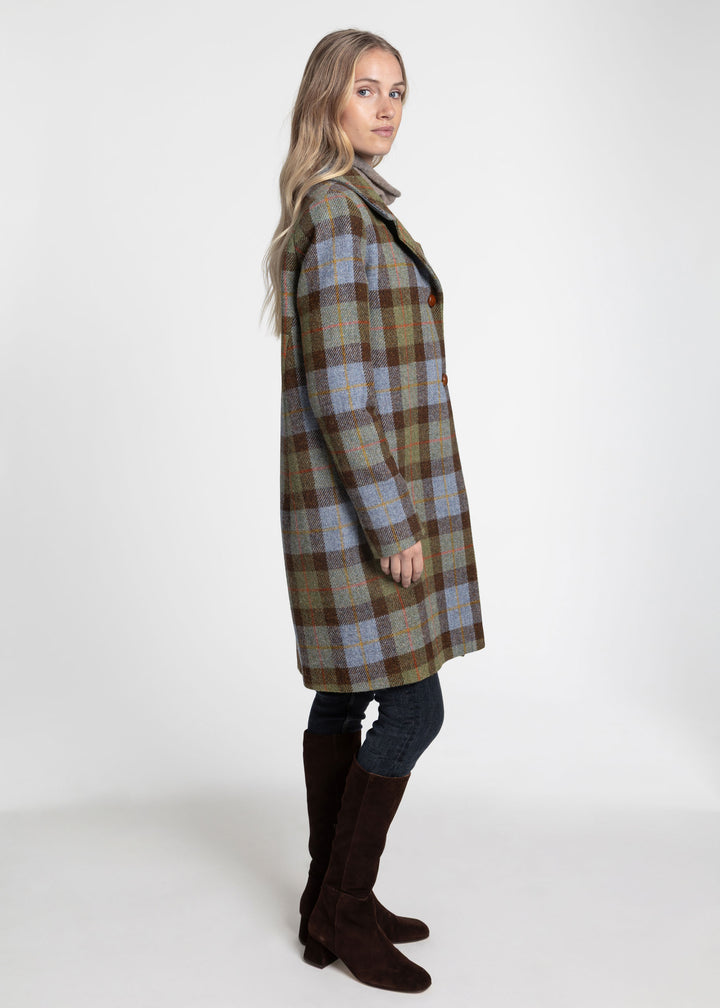 A woman modeling a knee-length plaid coat in side profile against a plain light gray studio background. The coat has a muted plaid pattern with brown, green, beige, and light blue tones.