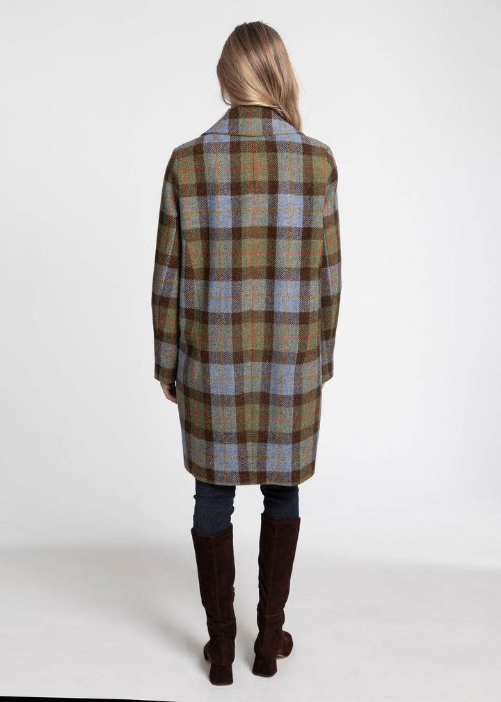 A woman viewed from behind wearing a mid-length plaid coat in brown, beige, and muted blue, paired with dark jeans and tall brown boots, against a plain white studio background.