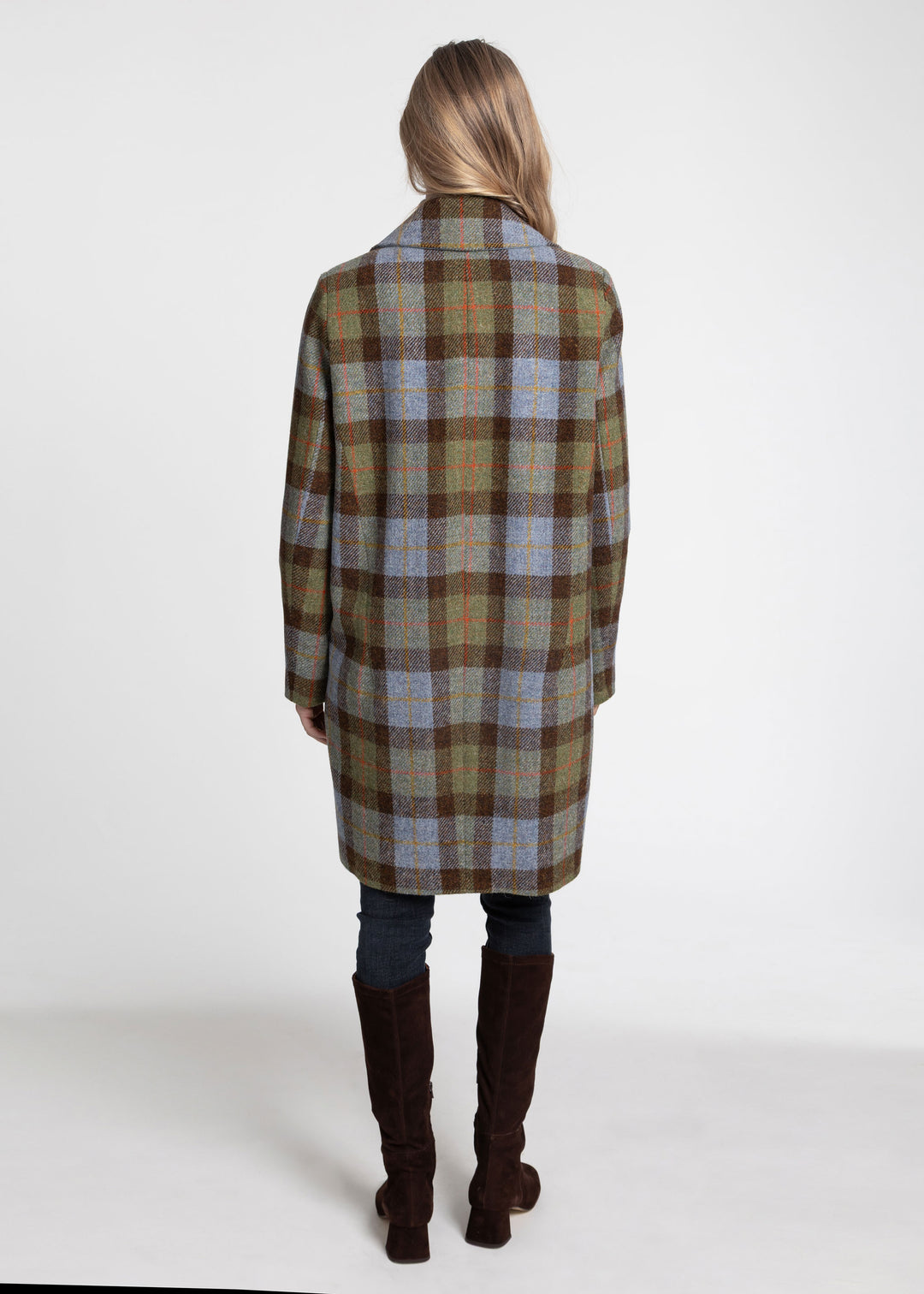 A woman viewed from behind wearing a mid-length plaid coat in brown, beige, and muted blue, paired with dark jeans and tall brown boots, against a plain white studio background.