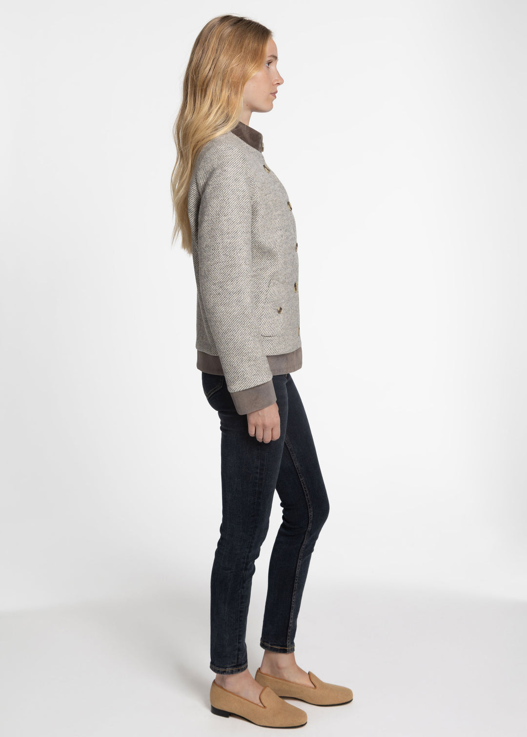 A woman shown in profile wearing a light gray textured jacket with brown trim, dark skinny jeans, and tan loafers against a plain white studio background.