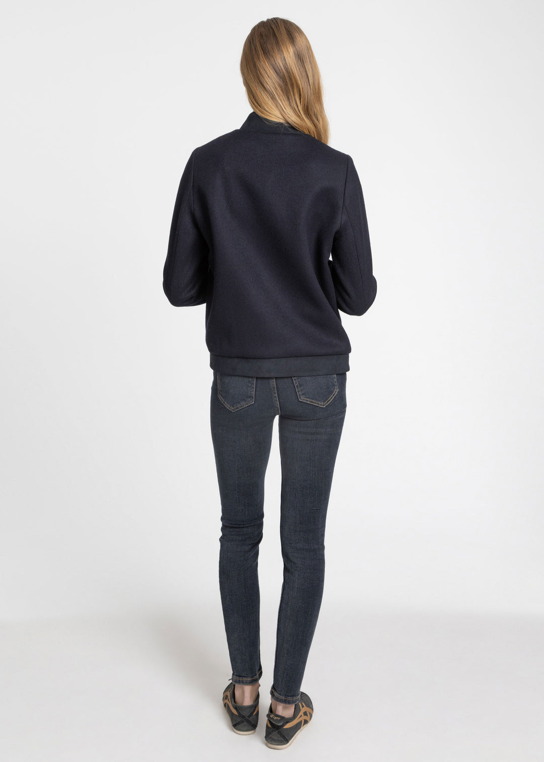 A woman viewed from behind wearing a dark navy blue jacket and dark gray skinny jeans standing against a plain white studio background.