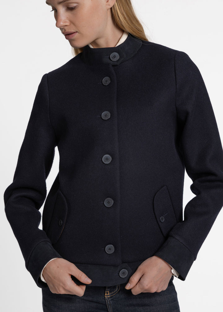 A woman wearing a dark navy blue wool-blend jacket with a high stand collar, dark buttons, and front flap pockets, shown from the chest down against a light gray studio background.