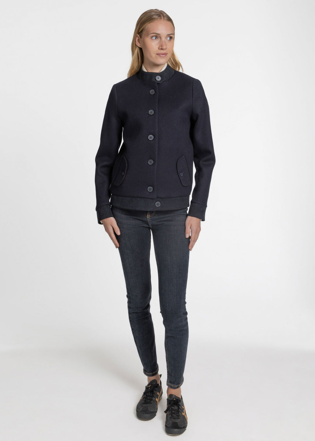 A woman modeling a dark navy blue wool-blend jacket with a high mock neck, button-front closure, and flap pockets, paired with dark gray skinny jeans and black sneakers, against a plain white studio background.