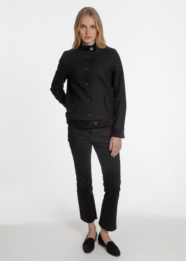 A woman modeling a black cropped jacket with a high collar and front buttons, paired with black pants and black slip-on shoes, against a plain white studio background.