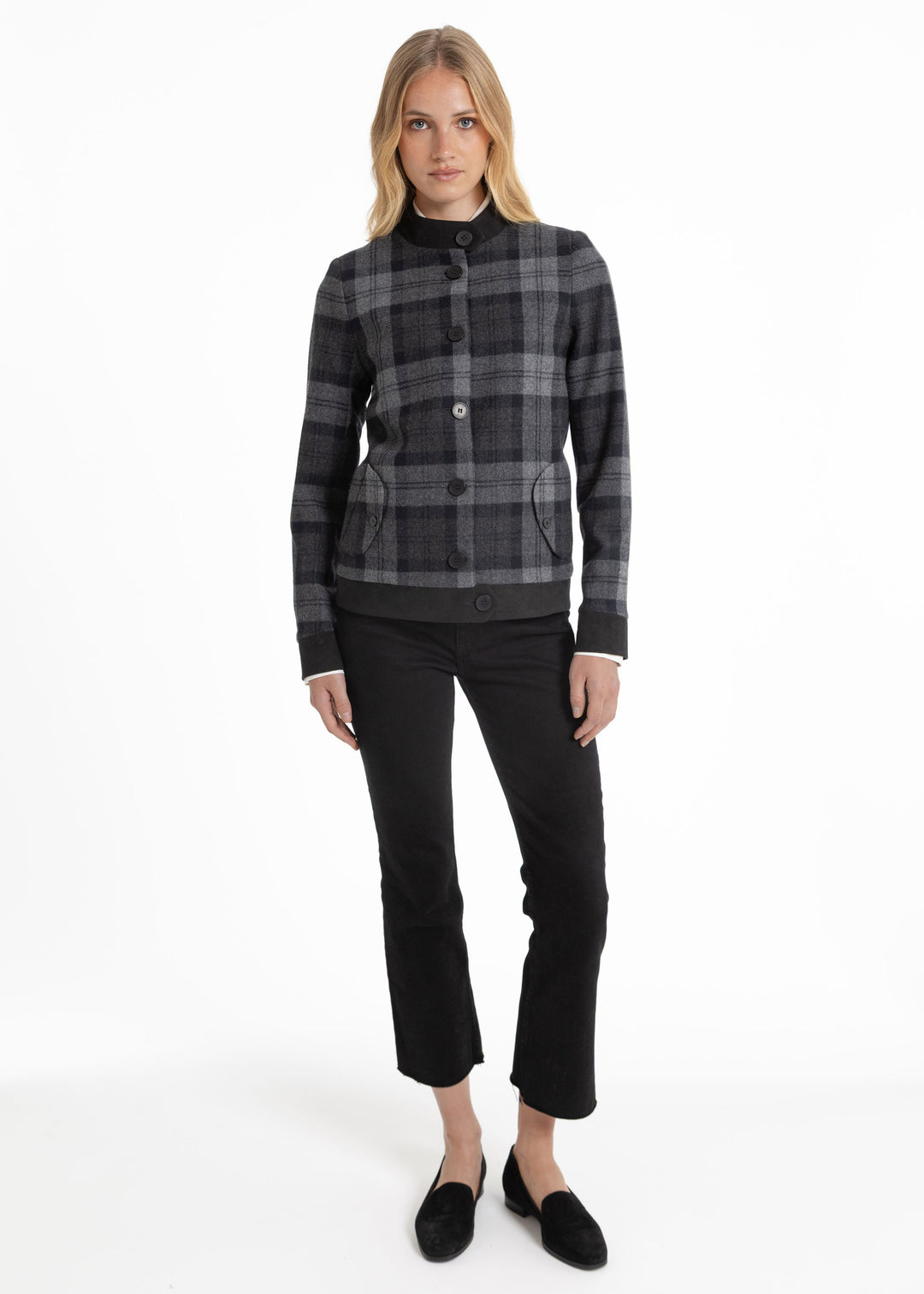 A woman modeling a black and gray plaid button-front jacket with a mock neck, paired with black cropped pants and black slip-on shoes, against a plain white studio background.