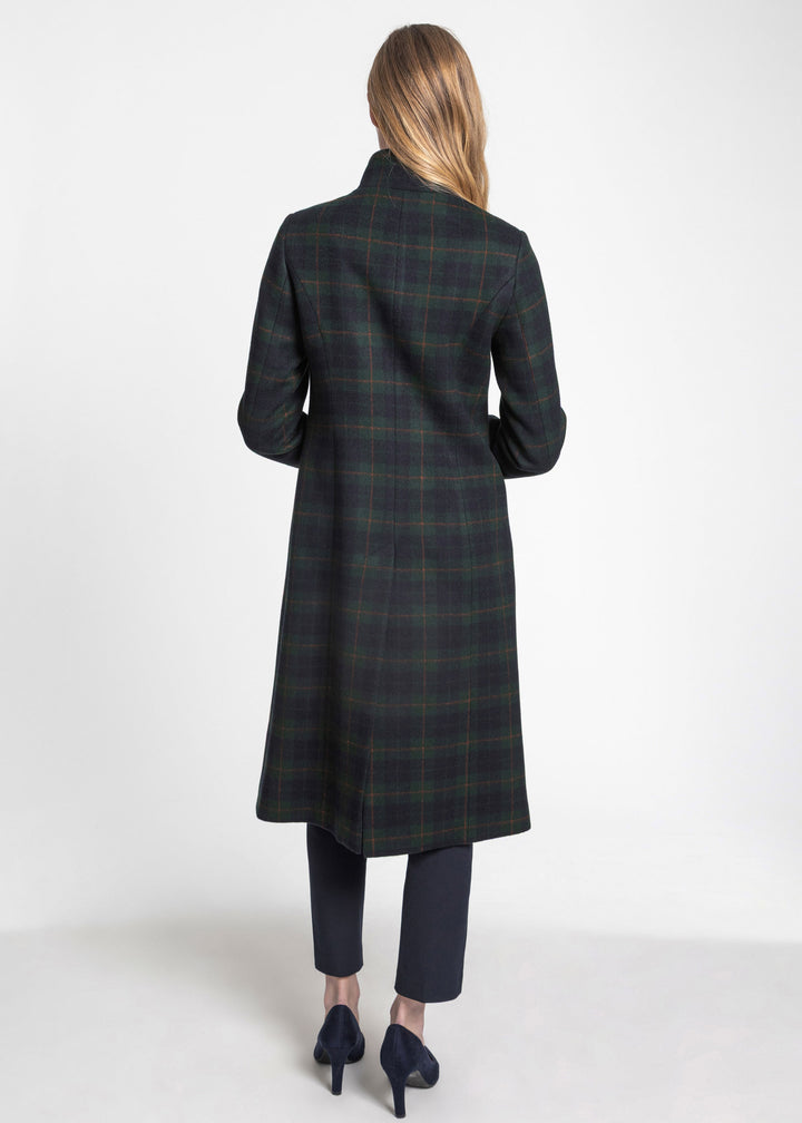 A woman viewed from the back wearing a long dark plaid coat, dark cropped trousers, and dark pointed heels against a plain light gray studio background.