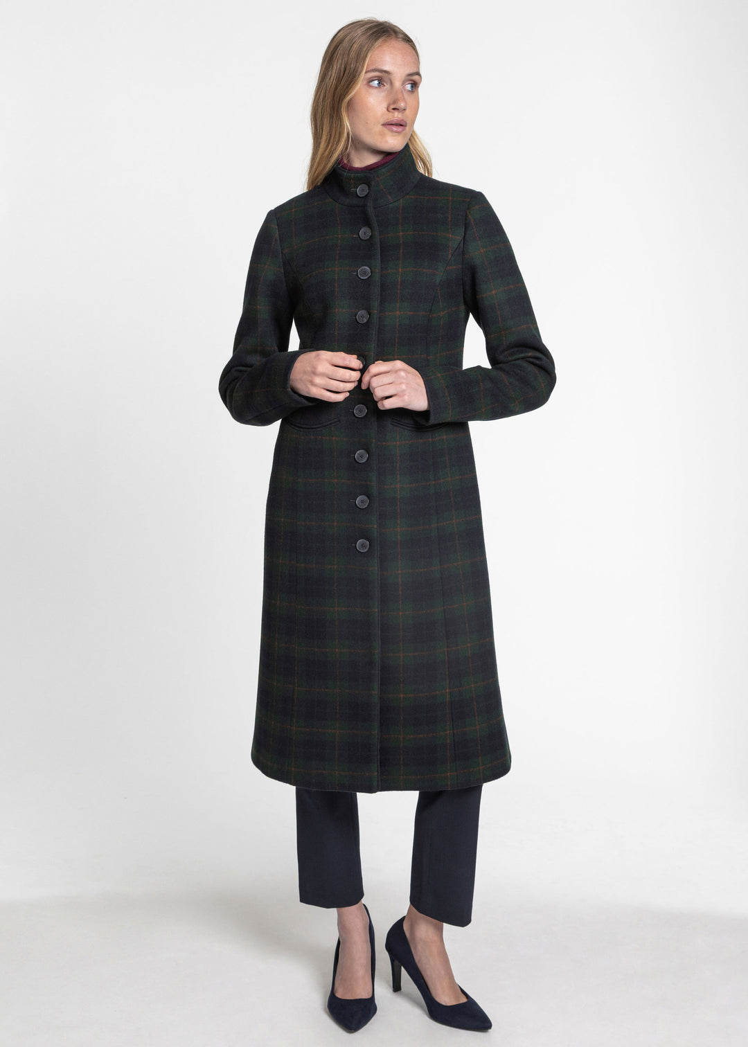 A woman modeling a long dark green and black plaid coat with a high collar and front buttons, paired with dark cropped trousers and black heels, against a plain white studio background.