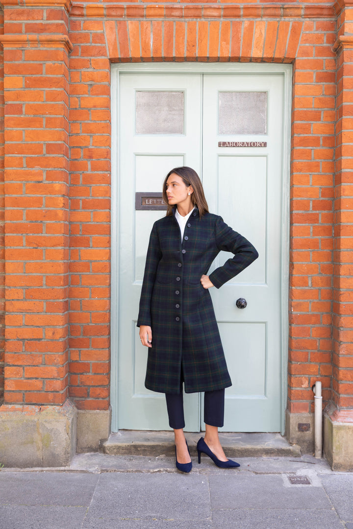 A woman stands in front of a light green door set in a red brick wall, wearing a long dark green and black plaid coat over a white top, dark cropped trousers, and navy heels.