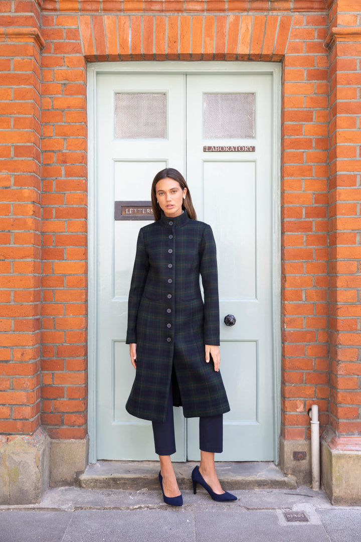 A woman stands in front of a light green door set in a red brick wall, wearing a long black and dark green plaid coat over a dark outfit with navy heels.