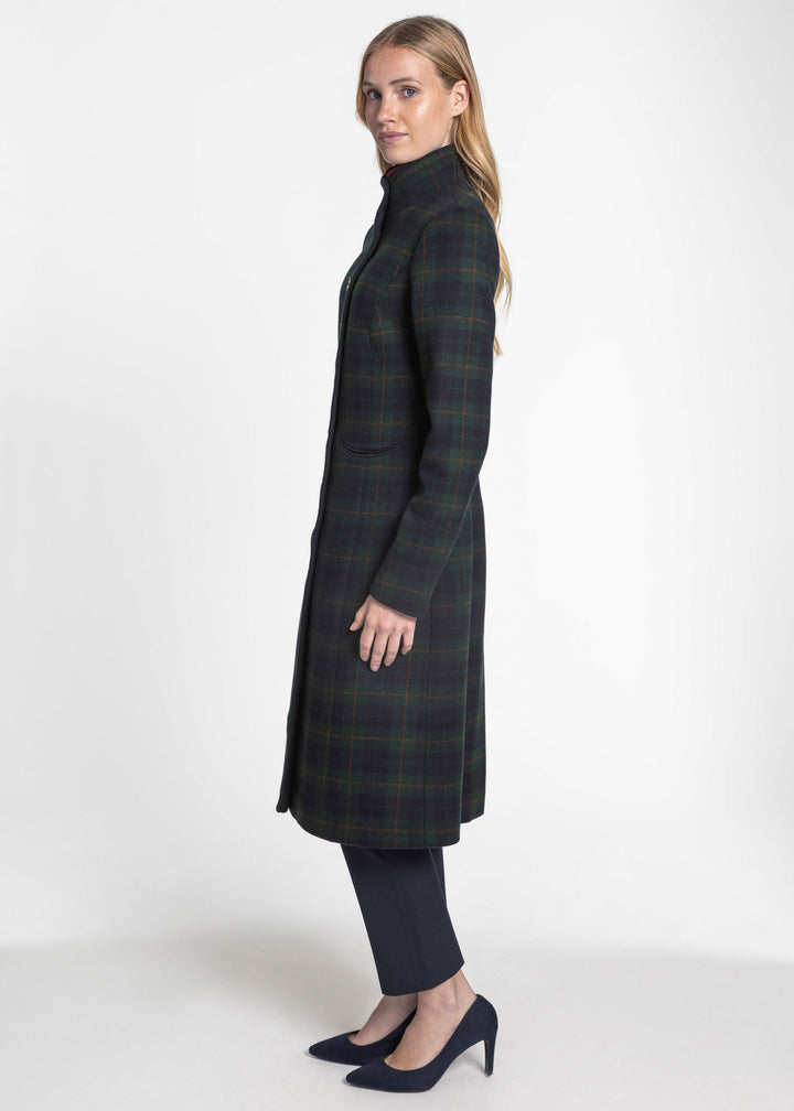A woman modeling a long dark green and black plaid coat with a high collar, paired with dark trousers and navy heels, photographed against a plain white studio background.