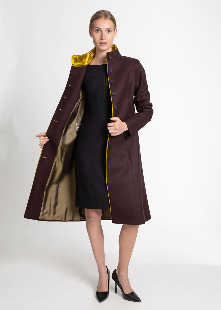A woman models a long dark mulberry coat with a shiny gold interior lining and a gold-trimmed collar, worn open over a black dress and black heels against a plain white studio background.