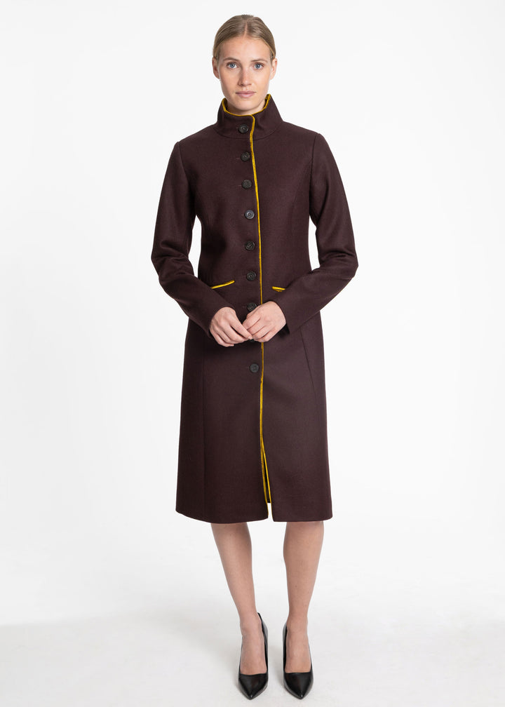 A woman modeling a dark mulberry knee-length coat with a high stand collar, front buttons, and contrasting gold piping details, set against a plain white studio background.