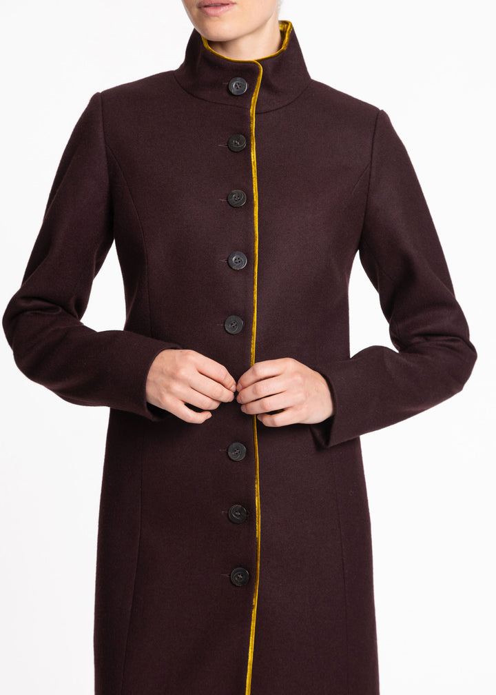 A model wearing a dark mulberry wool coat with a high stand collar, black buttons, and a contrasting yellow-gold trim running down the front.