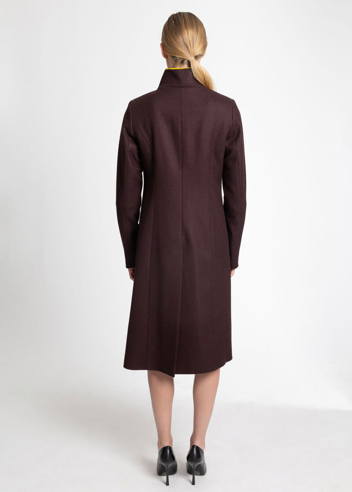 A woman viewed from the back wearing a long, dark mulberry coat with a high mock neck, paired with black pointed-toe heels, against a plain white studio background.