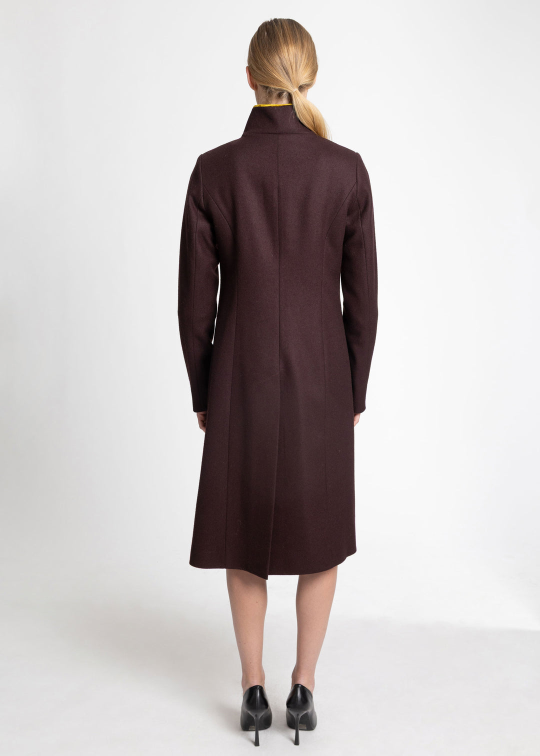 A woman viewed from the back wearing a long, dark mulberry coat with a high mock neck, paired with black pointed-toe heels, against a plain white studio background.
