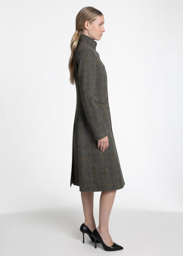 A woman in profile wearing a long, dark olive tweed coat with a high mock neck, paired with black heels, photographed against a clean white studio background.