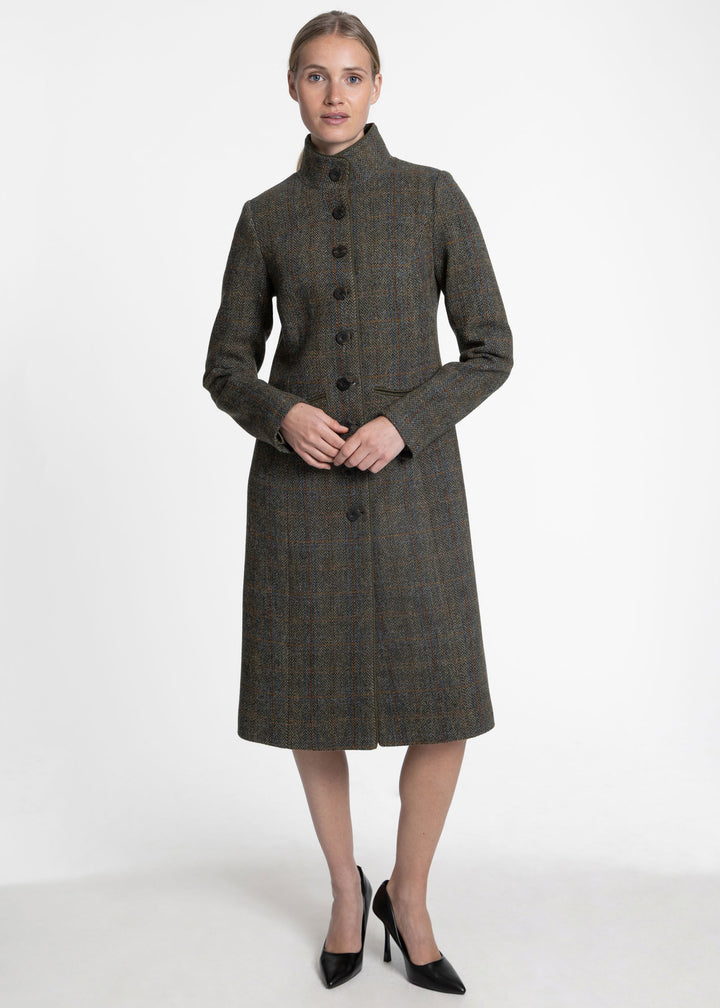 A woman modeling a knee-length high-neck green tweed coat with black buttons, and black high heels against a plain white studio background.