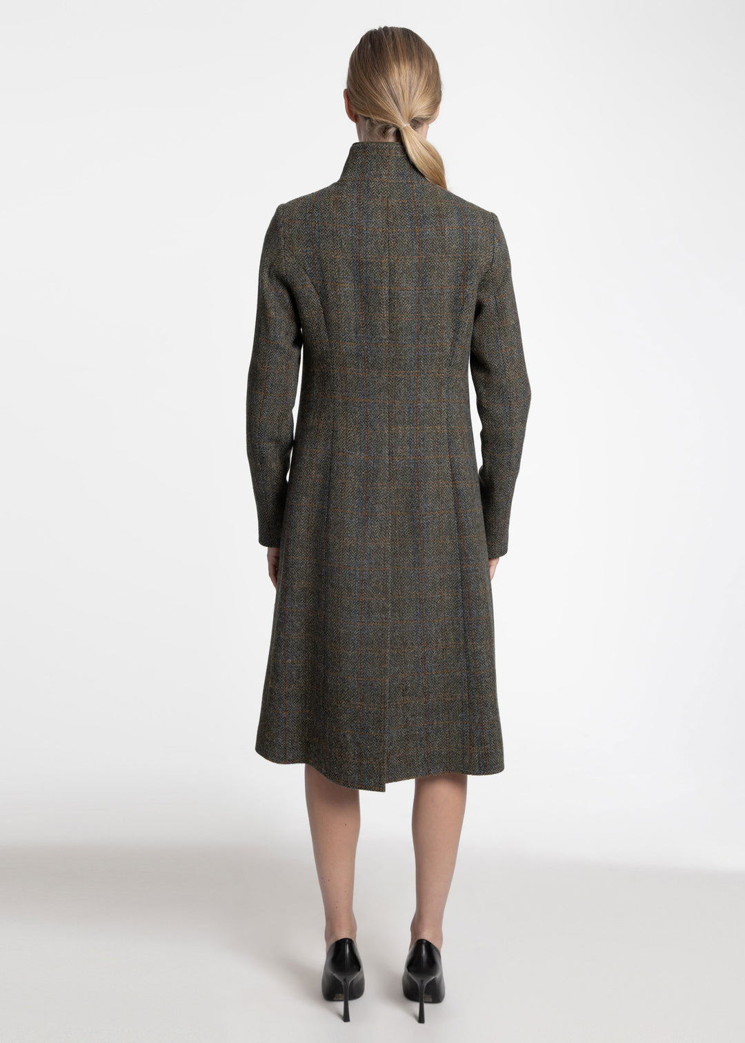 A woman viewed from the back wearing a long, knee-length plaid coat in dark olive green paired with black heels, against a plain white studio background.