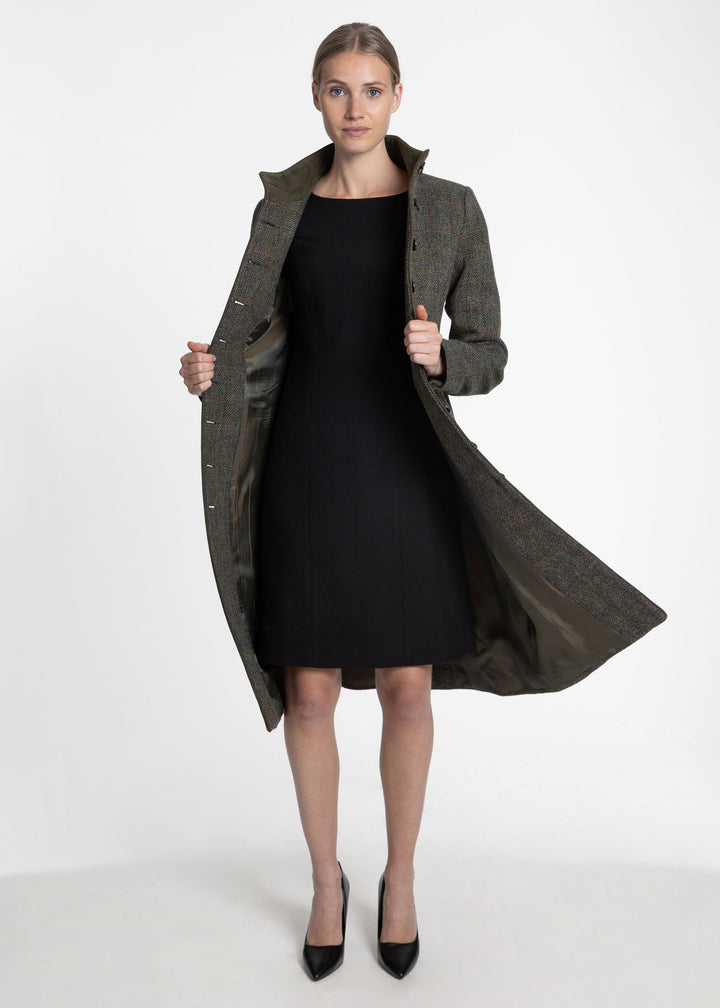 A woman modeling a long dark olive green patterned overcoat worn open over a black knee-length dress and black pointed heels, photographed against a clean light gray studio background.