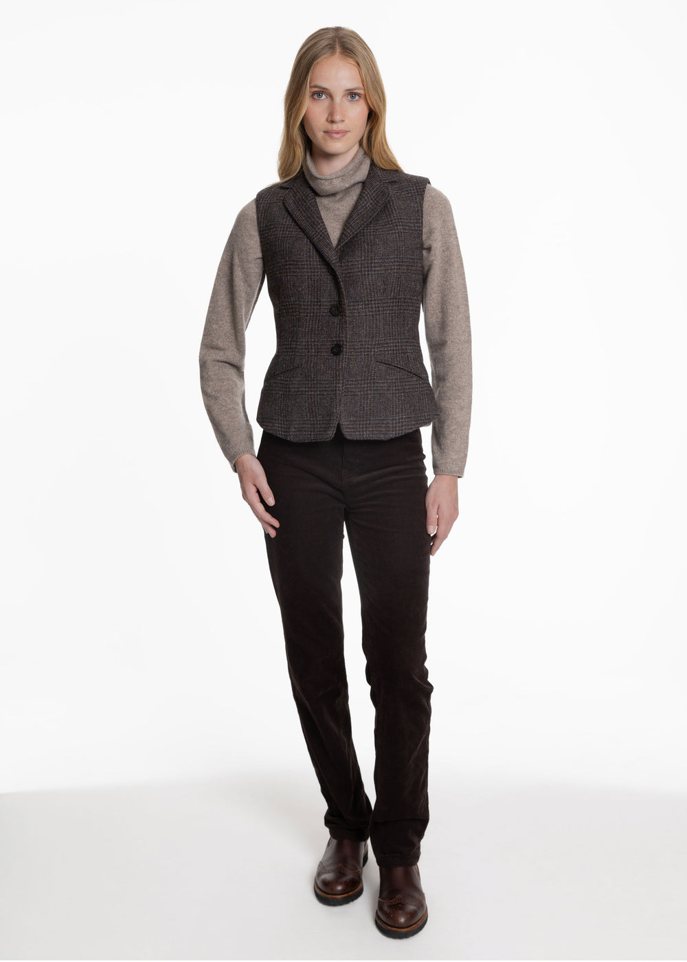 A woman wearing a black an brown check waistcoat over a grey turtleneck sweater. Worn with brown trousers and chelsea boots.